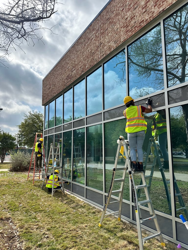 Workers in safety vests cleaning large glass windows of a brick building using ladders outdoors